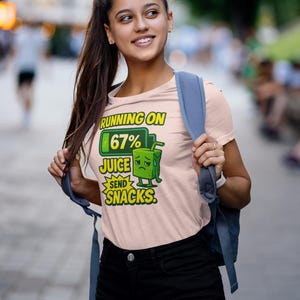Pu&ograve; includere: T-shirt rosa chiaro con la scritta "RUNNING ON 67% JUICE SEND SNACKS". La grafica presenta un personaggio di cartone di succo con una faccia triste. La persona indossa pantaloncini neri e uno zaino blu.