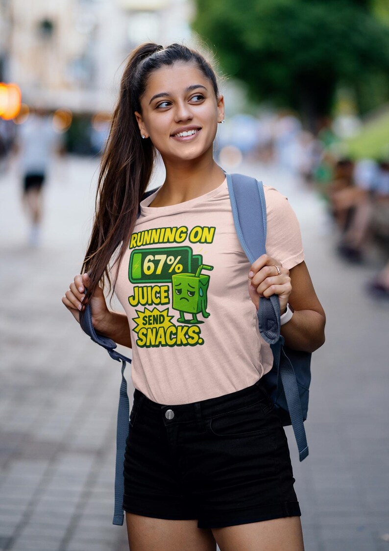 Pu&ograve; includere: T-shirt rosa chiaro con la scritta "RUNNING ON 67% JUICE SEND SNACKS". La grafica presenta un personaggio di cartone di succo con una faccia triste. La persona indossa pantaloncini neri e uno zaino blu.