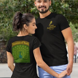 May include: Two people wearing black t-shirts. The woman's shirt has a peacock design with the text "Fountain of Youth" on the back. The man's shirt has a feather design with the text "Saint Augustine est. 1965".