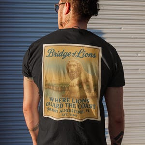 Może przedstawiać: Czarny t-shirt z grafiką w stylu vintage na plecach. Grafika przedstawia posąg lwa na moście z napisem "Bridge of Lions" i "Where Lions Guard the Coast, Saint Augustine, FL, EST. 1565."