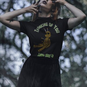 May include: Black t-shirt with the text "Thinking of You" and "Love Hurts" in green. The shirt features a voodoo doll graphic with pins. The person is wearing a black skirt and has dark nail polish.