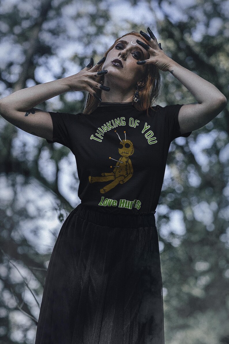 May include: Black t-shirt with the text "Thinking of You" and "Love Hurts" in green. The shirt features a voodoo doll graphic with pins. The person is wearing a black skirt and has dark nail polish.