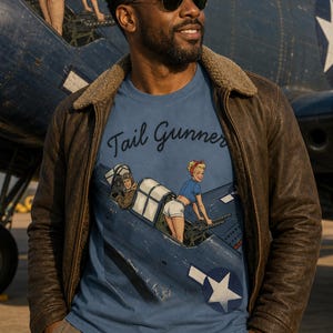 May include: A blue t-shirt featuring a graphic illustration of a vintage military aircraft with a pilot and a person operating a tail gun. The text "Tail Gunner" is printed in a dark script font above the aircraft illustration on the front.