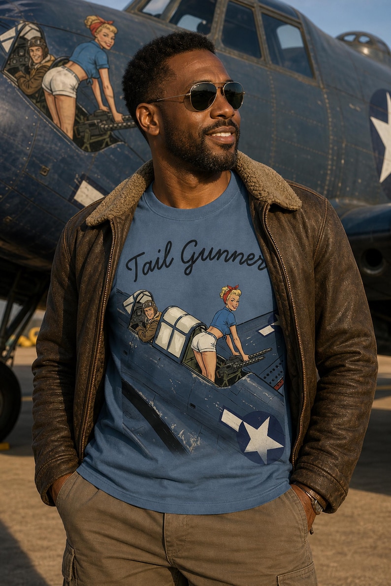 May include: A blue t-shirt featuring a graphic illustration of a vintage military aircraft with a pilot and a person operating a tail gun. The text "Tail Gunner" is printed in a dark script font above the aircraft illustration on the front.