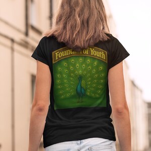 May include: Black t-shirt with a peacock design on the back. The peacock is teal with a green background and a banner that reads "Fountain of Youth". The shirt is worn with light blue jeans.