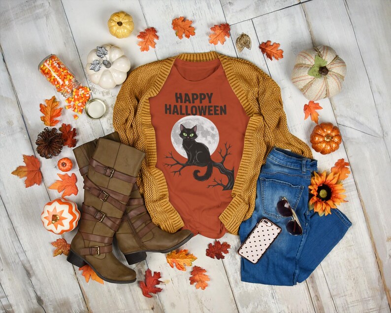 Może przedstawiać: Układ w stylu flat lay o tematyce Halloween z pomarańczową koszulką z grafiką czarnego kota i napisem "Happy Halloween". Zestaw zawiera musztardowy kardigan, niebieskie dżinsy, brązowe buty i dekoracyjne dynie i liście.