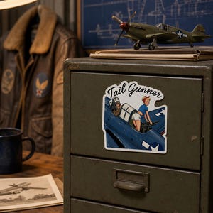 May include: A die-cut sticker featuring a pin-up style illustration of a person in a blue shirt and white shorts operating a machine gun in the cockpit of a blue military aircraft, with the text Tail Gunner written in a black cursive script above.