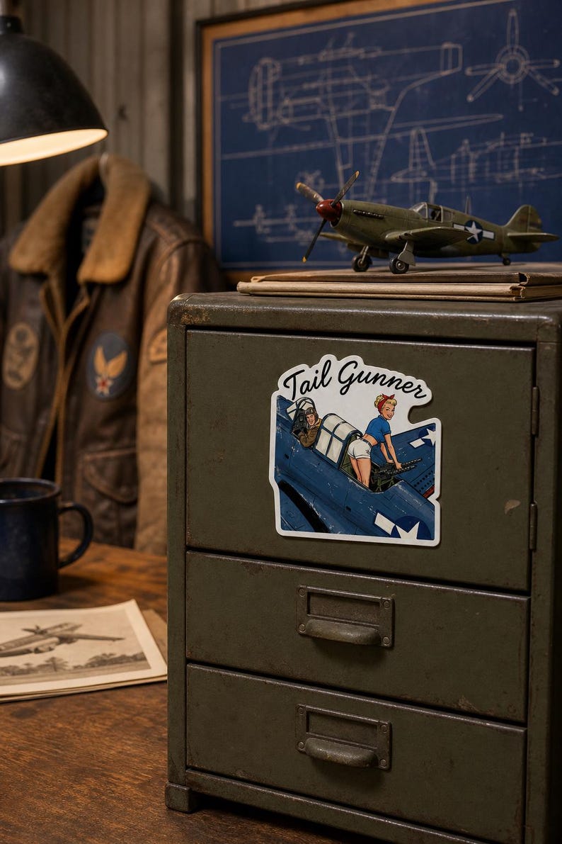 May include: A die-cut sticker featuring a pin-up style illustration of a person in a blue shirt and white shorts operating a machine gun in the cockpit of a blue military aircraft, with the text Tail Gunner written in a black cursive script above.
