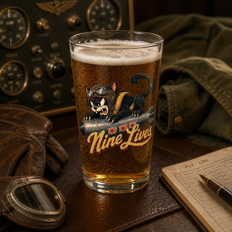 Op de afbeelding: Een helder glazen pint gevuld met goudkleurig bier en wit schuim, voorzien van een afbeelding van een zwarte kat met een vliegeniersbril die op een bom rijdt. De tekst Nine Lives staat in een geel lettertype onder de illustratie.