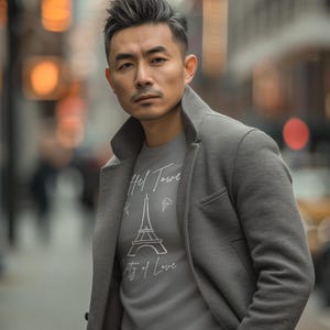 Op de afbeelding: Een persoon draagt een grijze jas over een grijs t-shirt met de Eiffeltoren en de tekst "Hel Tower City of Love". De achtergrond is een vage stadsstraat.
