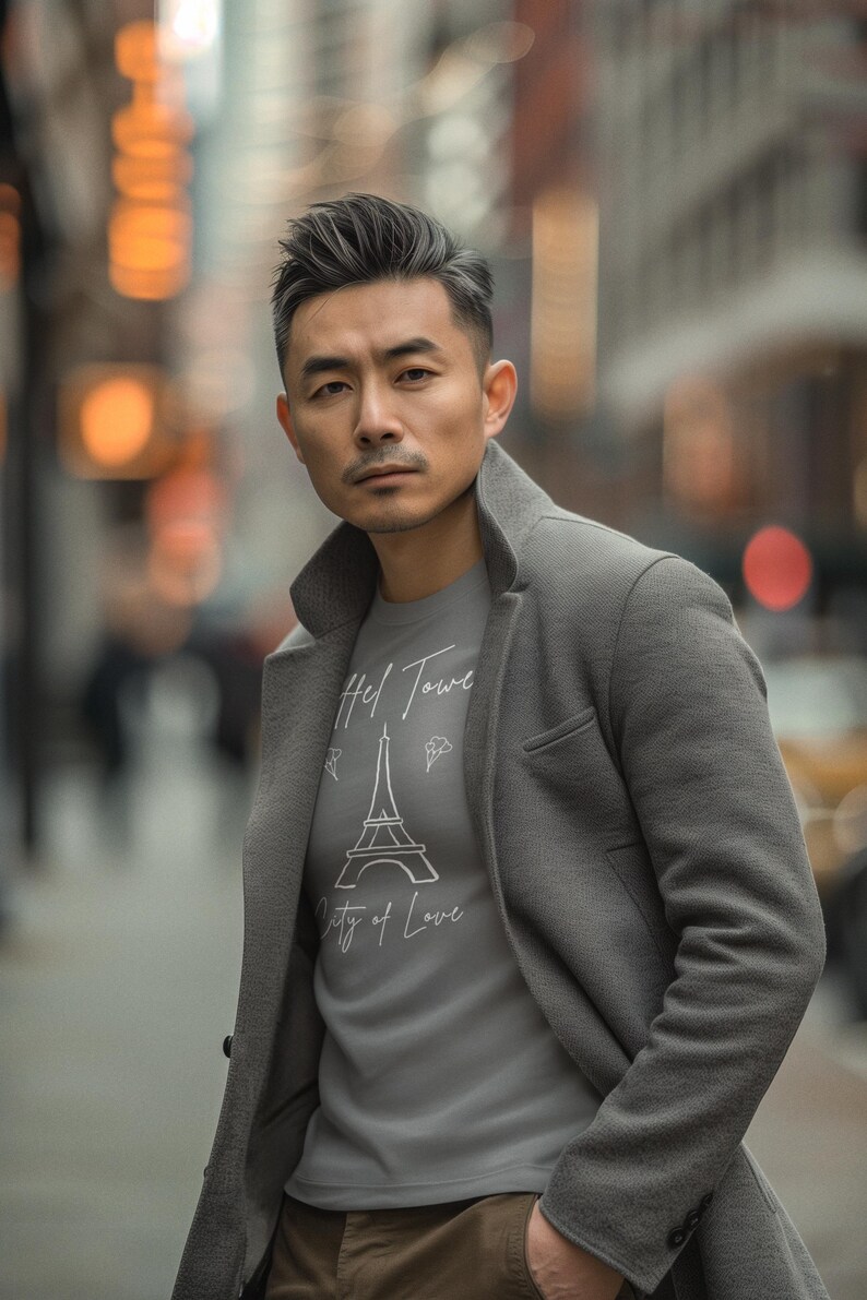 Op de afbeelding: Een persoon draagt een grijze jas over een grijs t-shirt met de Eiffeltoren en de tekst "Hel Tower City of Love". De achtergrond is een vage stadsstraat.