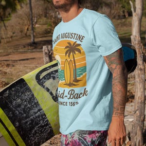 May include: Light blue t-shirt with a graphic featuring a sunset, palm tree, and surfboards, with the text "Saint Augustine" and "Mid-Back Since 1565." The person is wearing sunglasses and tropical print shorts, carrying a surfboard.