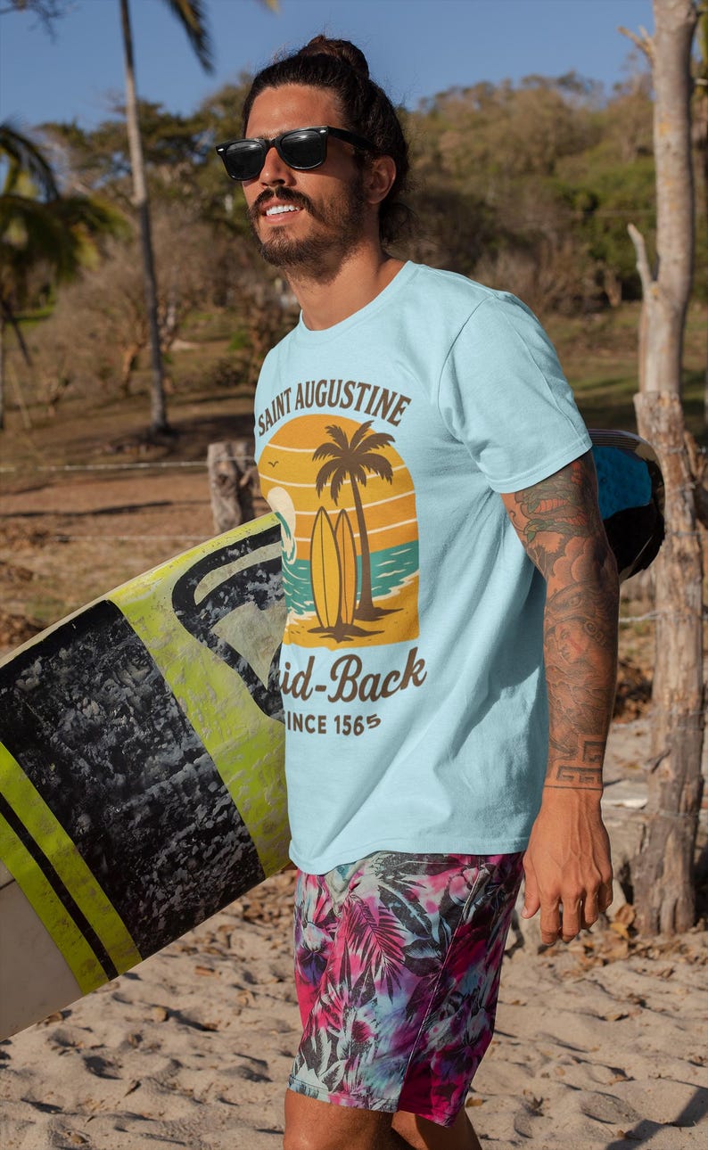 May include: Light blue t-shirt with a graphic featuring a sunset, palm tree, and surfboards, with the text "Saint Augustine" and "Mid-Back Since 1565." The person is wearing sunglasses and tropical print shorts, carrying a surfboard.