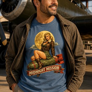 Peut inclure: Un t-shirt bleu avec une impression graphique repr&eacute;sentant une femme blonde en veste en cuir marron et jupe verte assise sur une bombe verte devant une grande lune jaune. Sous la bombe, une banni&egrave;re rouge porte le texte MIDNIGHT MISSION.