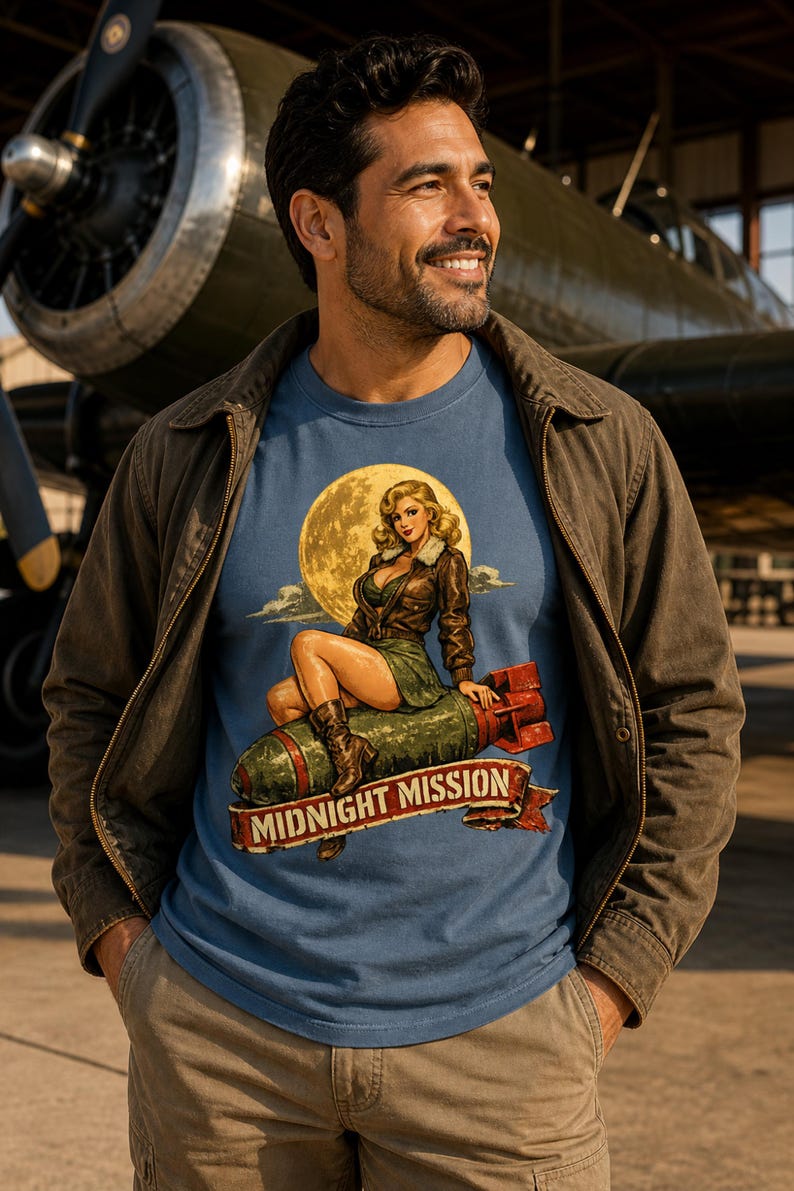 Peut inclure: Un t-shirt bleu avec une impression graphique repr&eacute;sentant une femme blonde en veste en cuir marron et jupe verte assise sur une bombe verte devant une grande lune jaune. Sous la bombe, une banni&egrave;re rouge porte le texte MIDNIGHT MISSION.