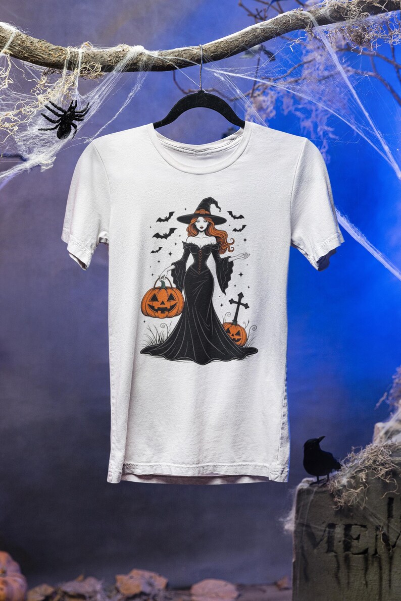 Może przedstawiać: Biała koszulka z ilustracją o tematyce Halloween. Grafika przedstawia wiedźmę w czarnej sukni i kapeluszu, trzymającą dynię. Nietoperze i gwiazdy otaczają wiedźmę, z krzyżem i dynią w tle.