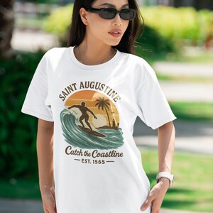 May include: White t-shirt featuring a graphic of a surfer riding a wave with a sunset backdrop. The text reads "Saint Augustine" and "Catch the Coastline - EST. 1565". The shirt is a casual wear item.