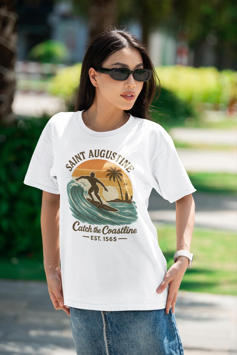 May include: White t-shirt featuring a graphic of a surfer riding a wave with a sunset backdrop. The text reads "Saint Augustine" and "Catch the Coastline - EST. 1565". The shirt is a casual wear item.