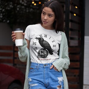 May include: White t-shirt featuring a black raven perched atop a skull, set against a clock face design. The shirt includes the text "Memento Mori MMXV". The model is wearing blue denim shorts and a light green cardigan.