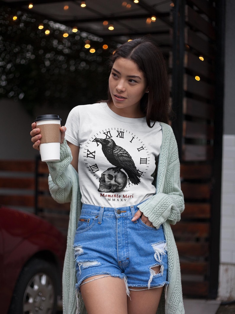 May include: White t-shirt featuring a black raven perched atop a skull, set against a clock face design. The shirt includes the text "Memento Mori MMXV". The model is wearing blue denim shorts and a light green cardigan.