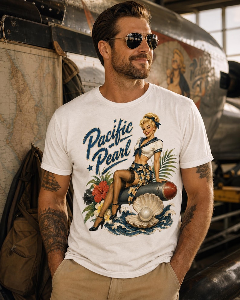 May include: A white short-sleeve t-shirt featuring a graphic print of a pin-up style woman sitting on a bomb, with the text Pacific Pearl, a large open seashell with a pearl, tropical flowers, and ocean waves in blue, red, and green tones.