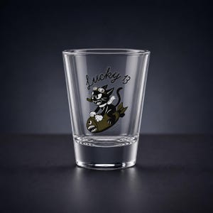 May include: A clear glass shot glass featuring a black and white graphic of a cartoon cat wearing goggles and a scarf while riding a bomb. The text Lucky 13 is printed in a cursive black font above the cat illustration on the front of the glass.