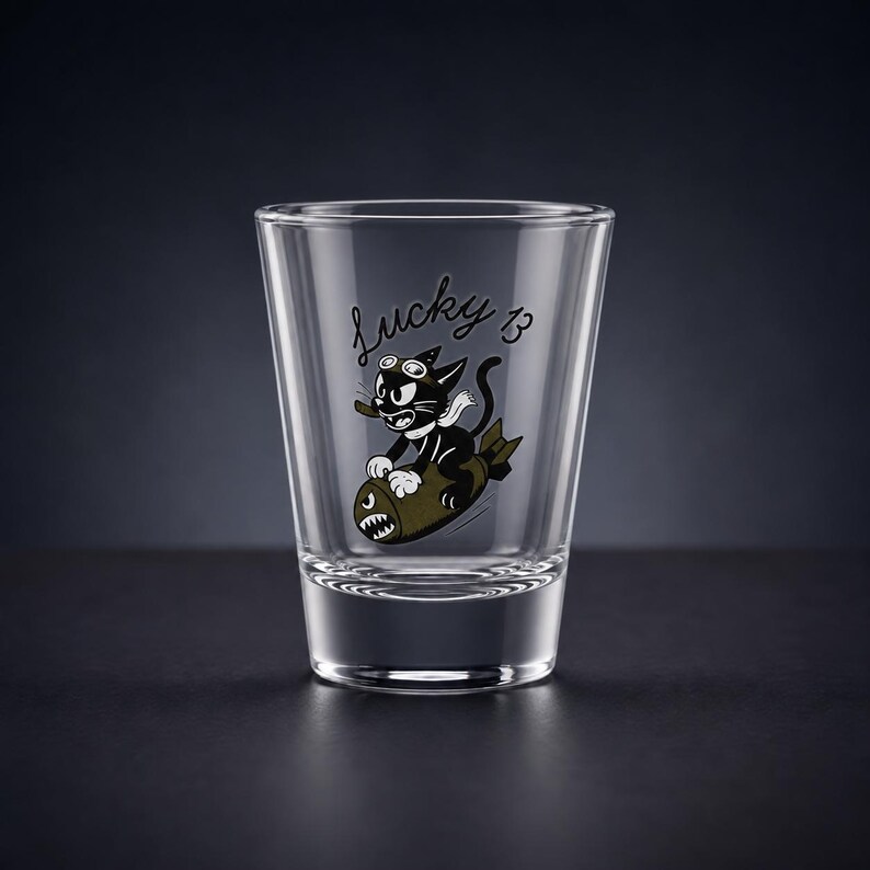 May include: A clear glass shot glass featuring a black and white graphic of a cartoon cat wearing goggles and a scarf while riding a bomb. The text Lucky 13 is printed in a cursive black font above the cat illustration on the front of the glass.