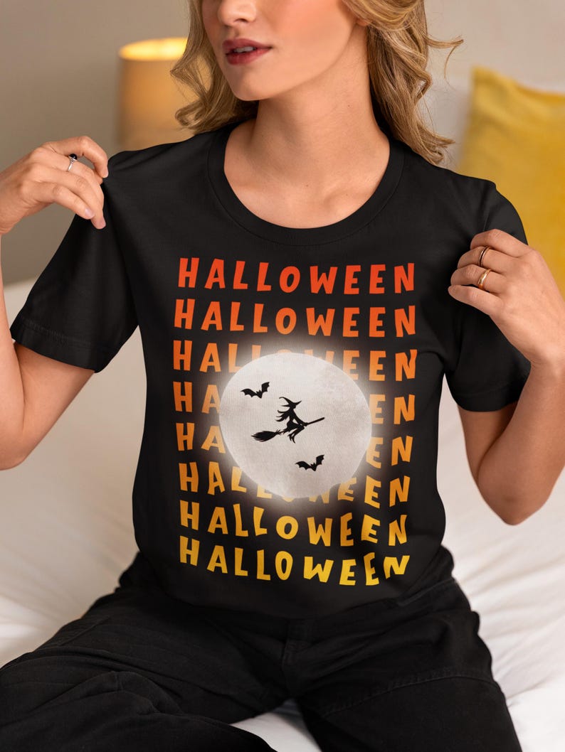 May include: Black t-shirt with the word "HALLOWEEN" in orange and yellow gradient text. A white full moon graphic features a witch on a broom and bats. The shirt is a crew neck style.
