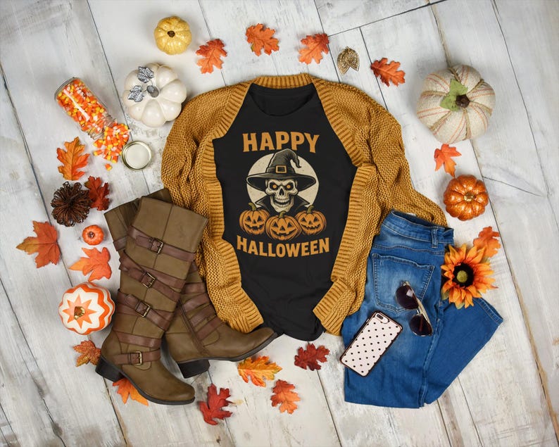 May include: A flat lay featuring a black t-shirt with a skull wearing a witch hat and the words "Happy Halloween". The shirt is paired with a mustard yellow cardigan, blue jeans, and brown boots. Decorative pumpkins and fall leaves surround the items.