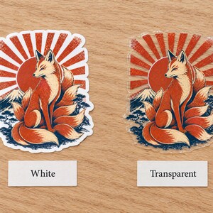 May include: Two stickers featuring a nine-tailed fox sitting before a red sunburst, a mountain, and trees. One sticker has a white border and backing, while the other is transparent. Labels below the stickers read White and Transparent.