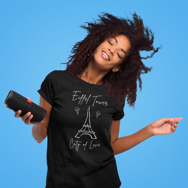Op de afbeelding: Zwart T-shirt met de afbeelding van de Eiffeltoren en de tekst "Eiffel Tower City of Love". Een zwarte cilindrische luidspreker wordt in de linkerhand gehouden. De achtergrond is effen lichtblauw.