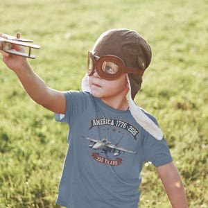 May include: A blue short-sleeved t-shirt featuring a graphic of a fighter jet with the text AMERICA 1776-2026 and 250 YEARS. The shirt is worn with a brown aviator cap with goggles and a small wooden toy airplane is held in the air.