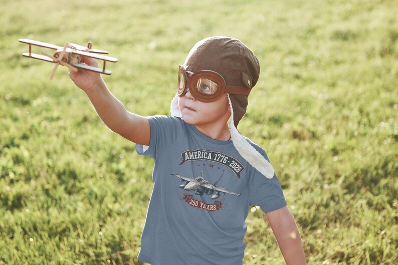 May include: A blue short-sleeved t-shirt featuring a graphic of a fighter jet with the text AMERICA 1776-2026 and 250 YEARS. The shirt is worn with a brown aviator cap with goggles and a small wooden toy airplane is held in the air.