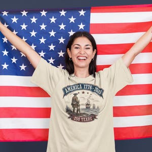 May include: A cream-coloured t-shirt featuring a graphic print with the text AMERICA 1776-2026 and 250 YEARS. The design depicts a historical figure standing on a shore looking toward ships at sea. The shirt is displayed against a large American flag.