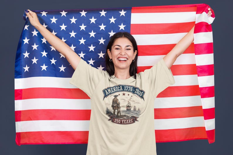 May include: A cream-coloured t-shirt featuring a graphic print with the text AMERICA 1776-2026 and 250 YEARS. The design depicts a historical figure standing on a shore looking toward ships at sea. The shirt is displayed against a large American flag.