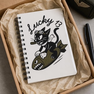 May include: A white spiral-bound notebook featuring a black and white illustration of a cartoon cat wearing goggles and a scarf while riding an olive green bomb. The text Lucky 13 is printed in black script above the cat. The notebook is inside a box.