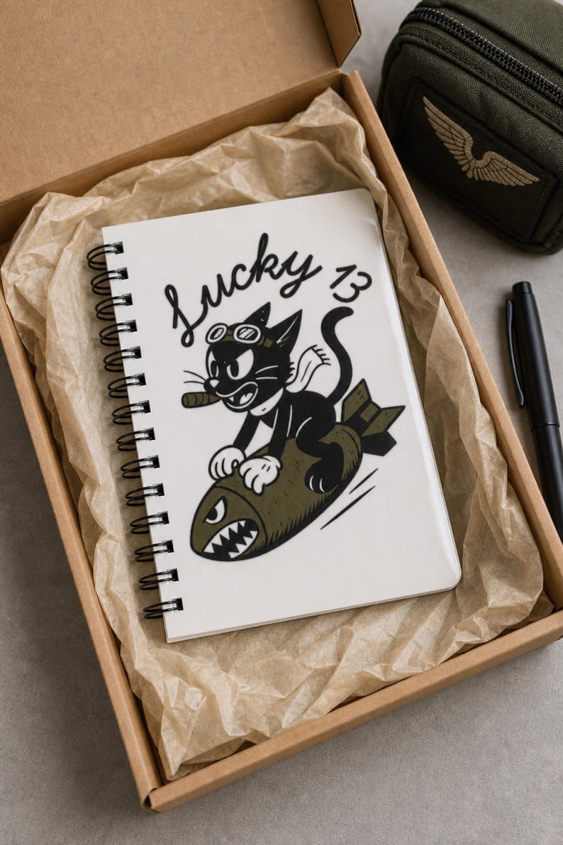 May include: A white spiral-bound notebook featuring a black and white illustration of a cartoon cat wearing goggles and a scarf while riding an olive green bomb. The text Lucky 13 is printed in black script above the cat. The notebook is inside a box.