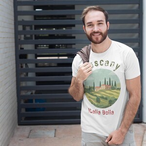 May include: A white t-shirt featuring a circular graphic of a Tuscan landscape with rolling hills, cypress trees, and a villa. The text "Tuscany" and "Italia Bella" are also on the shirt. The person is wearing gray shorts.