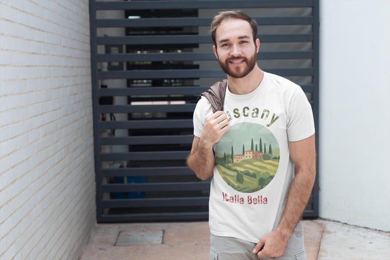 May include: A white t-shirt featuring a circular graphic of a Tuscan landscape with rolling hills, cypress trees, and a villa. The text "Tuscany" and "Italia Bella" are also on the shirt. The person is wearing gray shorts.