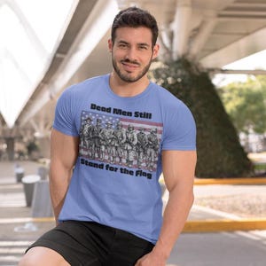 May include: A light blue short-sleeve t-shirt featuring a graphic of soldiers standing in front of an American flag. The text "Dead Men Still Stand for the Flag" is printed in black lettering above and below the central illustration.
