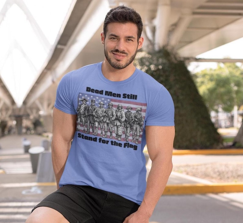 May include: A light blue short-sleeve t-shirt featuring a graphic of soldiers standing in front of an American flag. The text "Dead Men Still Stand for the Flag" is printed in black lettering above and below the central illustration.