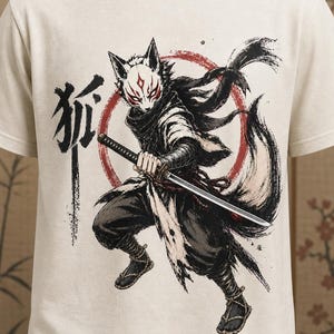 May include: A cream-colored t-shirt featuring a graphic illustration of a warrior wearing a fox mask and holding a katana. The design includes a large red circle, flowing dark clothing, and the Japanese kanji character for fox in black ink.