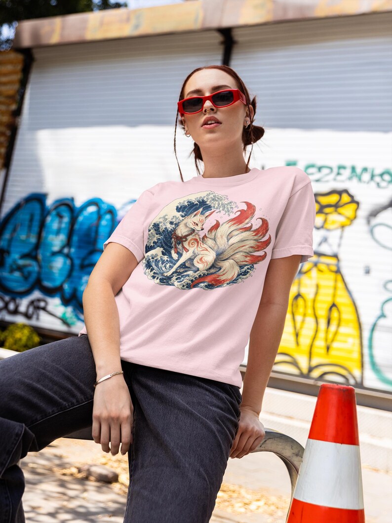 May include: A light pink short-sleeved t-shirt featuring a graphic print of a multi-tailed fox surrounded by stylized blue ocean waves. The fox has white and red fur with intricate markings and is positioned centrally on the chest of the garment.