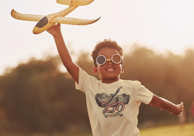 May include: A cream-colored short-sleeve t-shirt featuring a graphic print of Mount Rushmore with the text 250 and 1776-2020. The shirt is worn with round, dark-tinted goggles with a metallic frame, and a yellow foam toy airplane is held aloft.