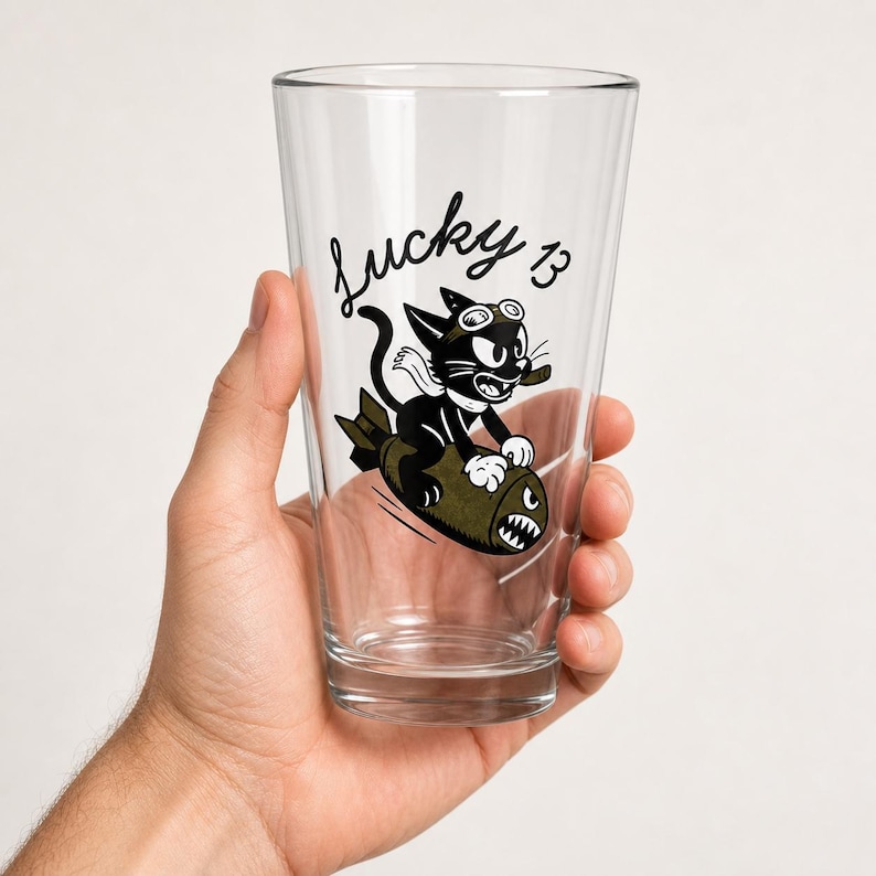 May include: A clear glass pint tumbler featuring a black graphic of a cartoon cat wearing goggles and riding an olive green bomb. The text Lucky 13 is printed in a black cursive script above the cat illustration on the front of the glass.