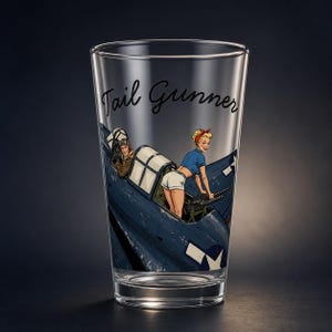 May include: A clear glass pint glass featuring a printed graphic of a blue military aircraft with a pilot in the cockpit and a person in a blue shirt and white shorts operating a machine gun. The text Tail Gunner is printed in black script.