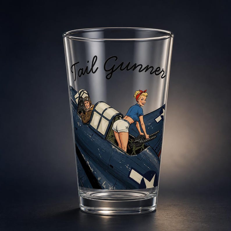 May include: A clear glass pint glass featuring a printed graphic of a blue military aircraft with a pilot in the cockpit and a person in a blue shirt and white shorts operating a machine gun. The text Tail Gunner is printed in black script.