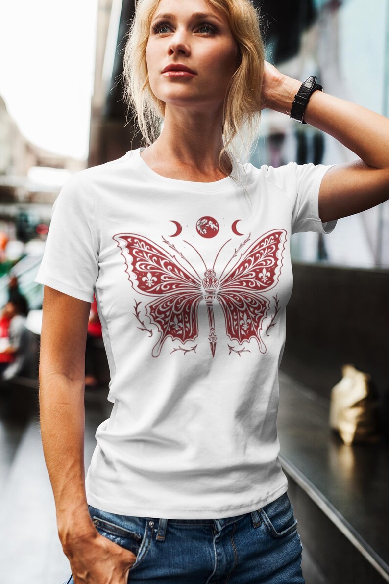 May include: White t-shirt featuring a detailed burgundy butterfly design with fleur-de-lis patterns and a central dagger. Above the butterfly are two crescent moons and a red globe. The shirt is a crew neck.