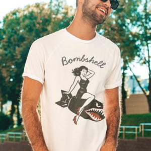 May include: White t-shirt with a vintage-style graphic of a woman riding a bomb, the word "Bombshell" above. The illustration features a woman in a black dress and red heels. The shirt is a casual, short-sleeved design.