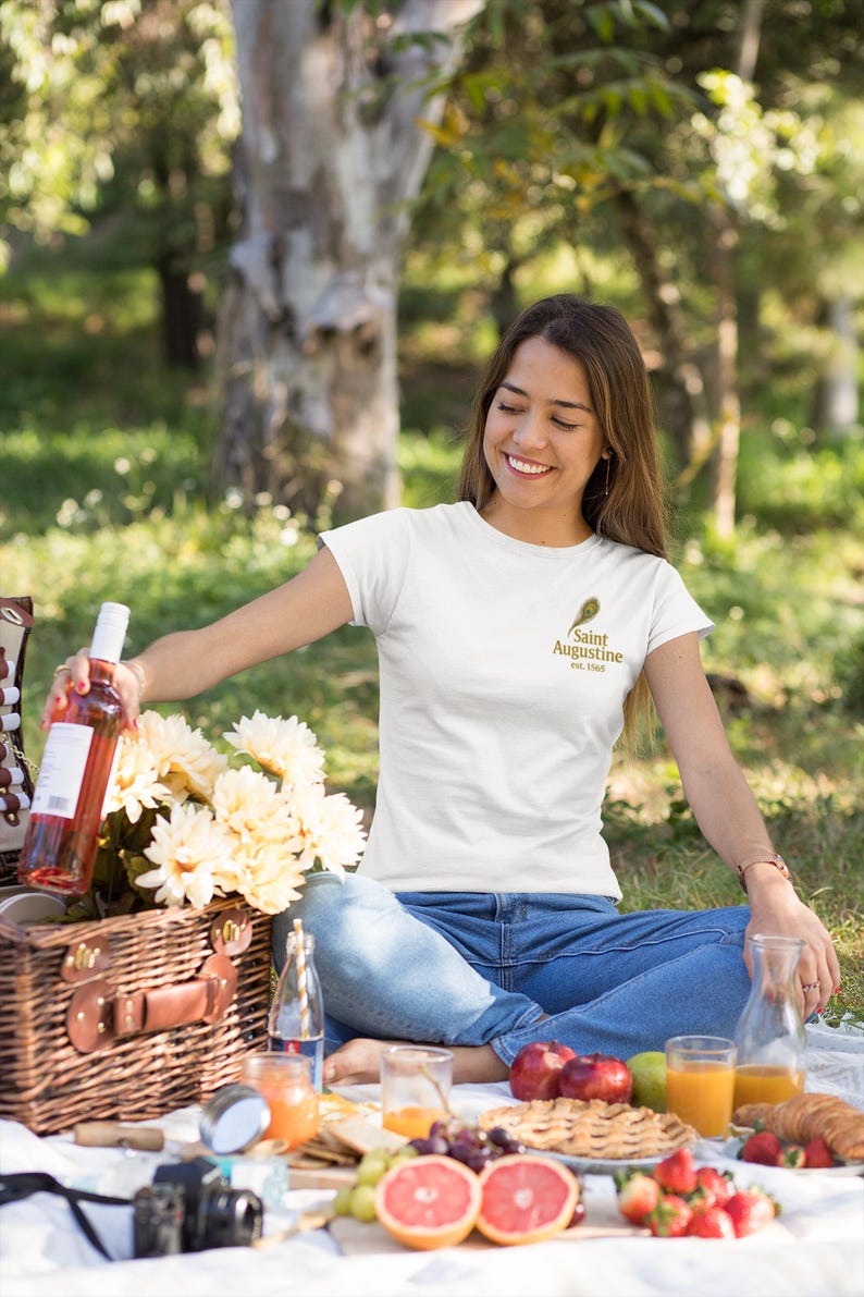 May include: A woman wearing a white t-shirt with a gold logo that says "Saint Augustine Est. 1565" is sitting on a picnic blanket. A picnic basket, wine bottle, flowers, and food are in front of her. The scene is outdoors.